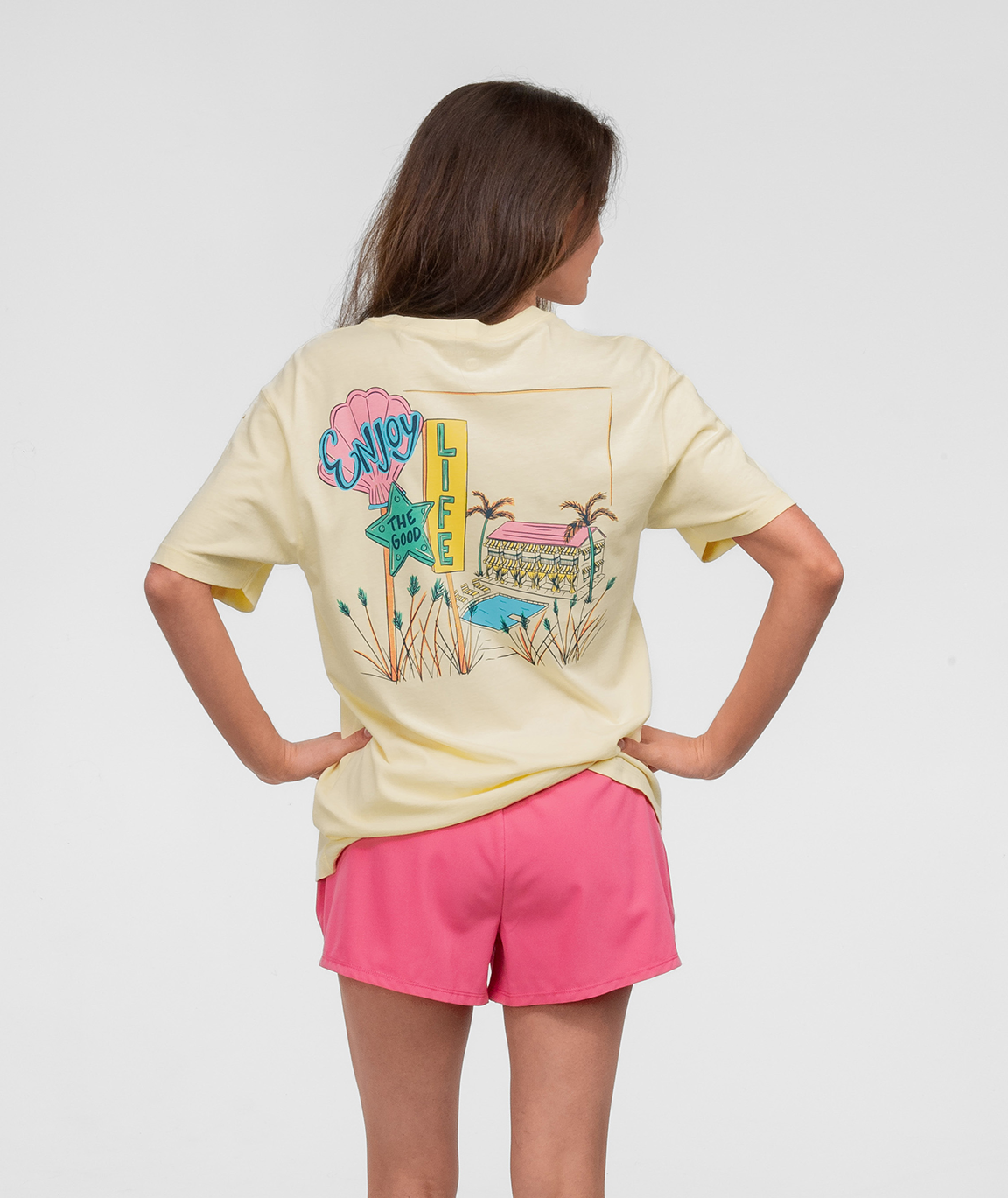 Girl wearing Hotel Good Life Tee SS with colorful graphic design on the back, paired with pink shorts.