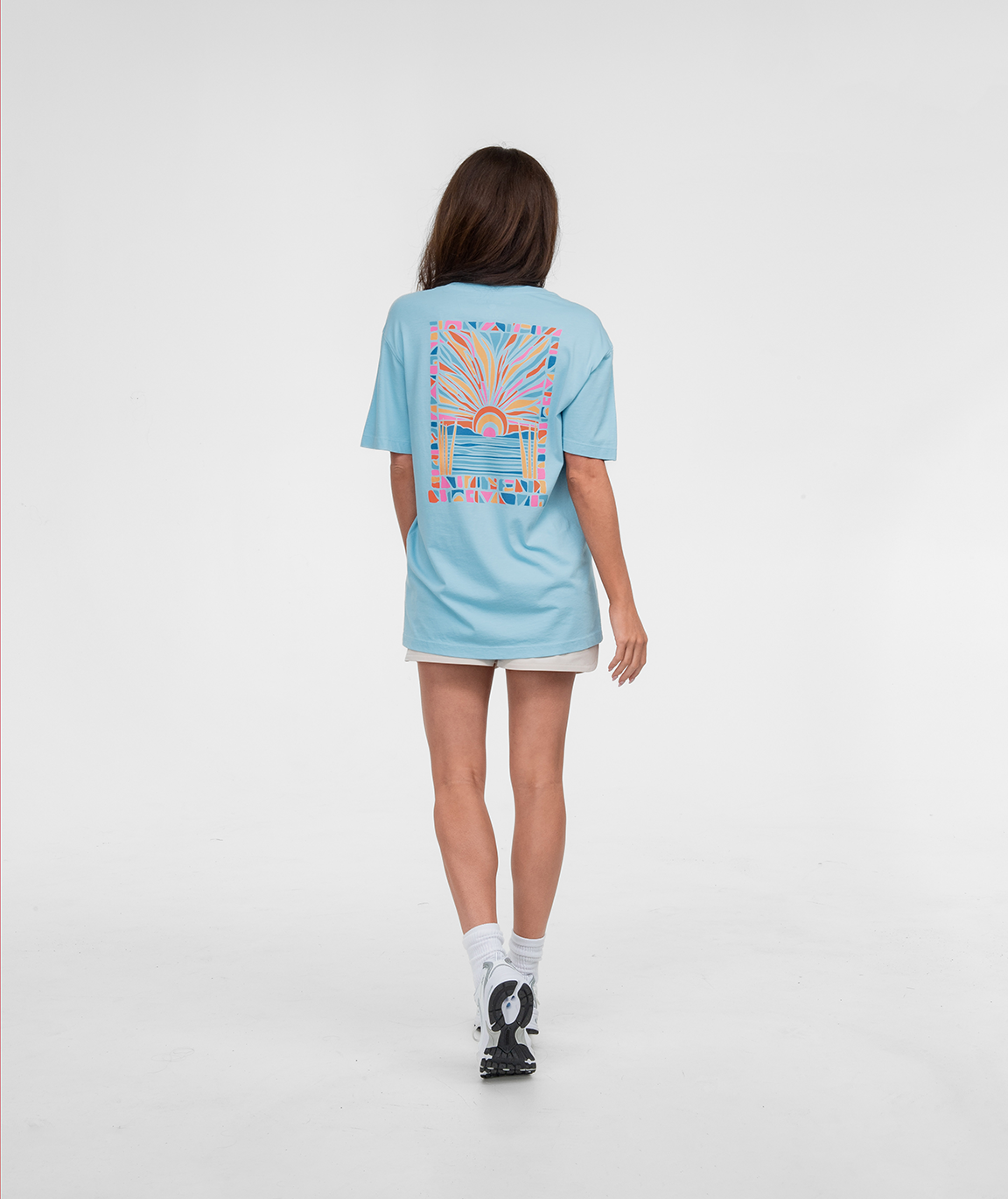 Woman wearing Pieces Of Paradise Tee SS from the back, featuring a colorful design on a light blue shirt.