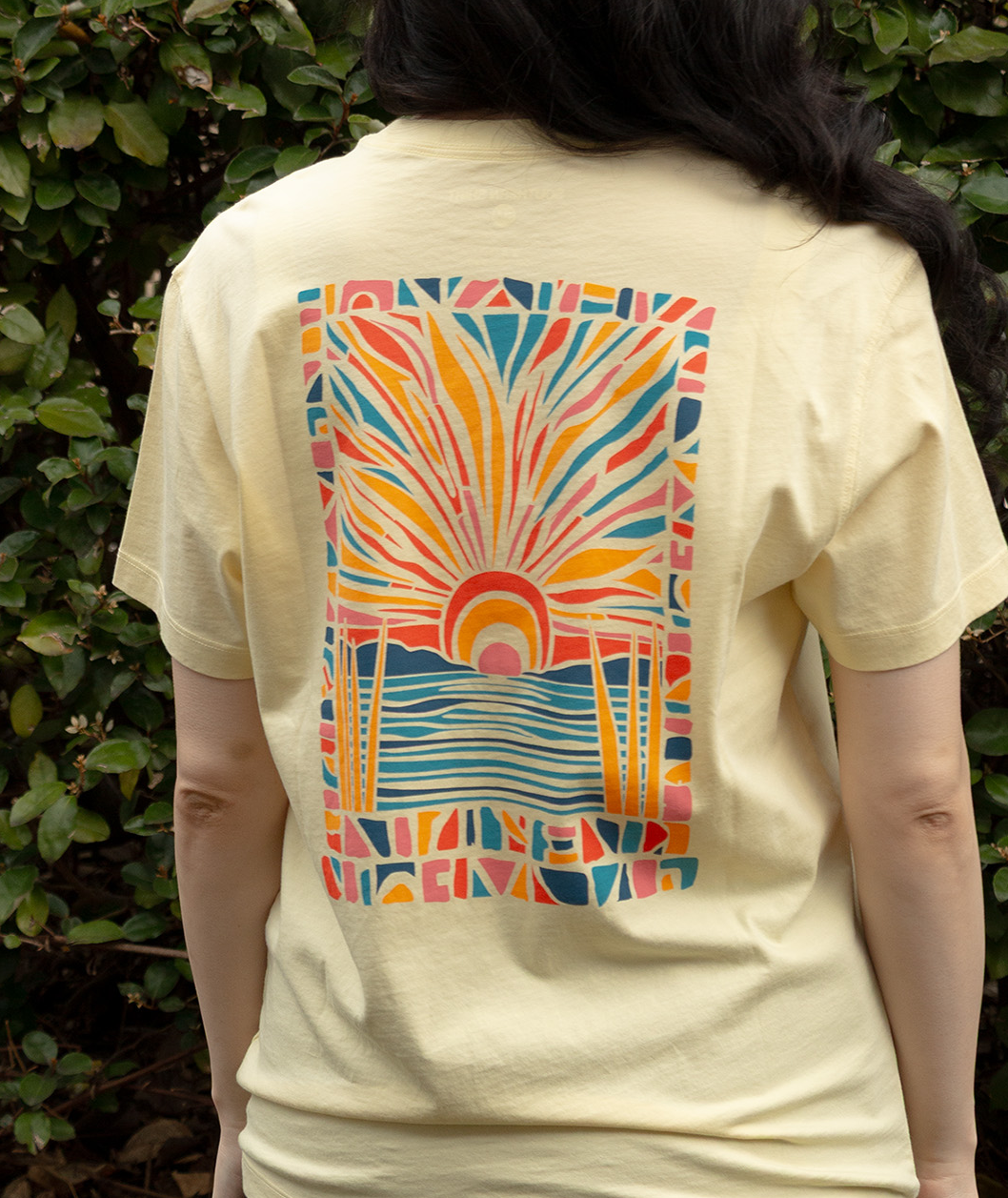 Woman wearing Pieces Of Paradise Tee SS with colorful sun and water design on the back against greenery.